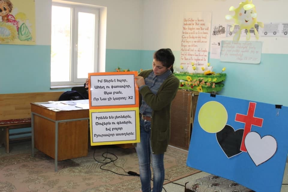 A teacher holds up a memory-verse poster beside a Wordless Book visual in a children's classroom